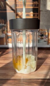 Milk is being poured into a glass blender cup containing egg yolks and egg whites, perfect for making a High Protein Dutch Baby, with a sunny kitchen and pastries visible in the blurred background.