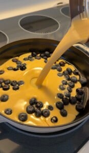 A creamy high protein Dutch baby batter is being poured into a pan full of fresh blueberries, preparing a dessert on a stovetop.