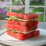 A stack of square watermelon slices layered with cheese and greens, held together by a toothpick, sits on a white plate with a sunlit garden visible through a window in the background.
