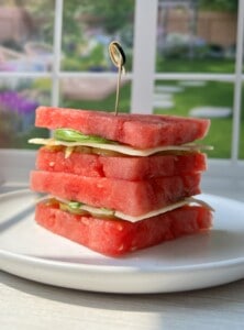 A stack of square watermelon slices layered with cheese and greens, held together by a toothpick, sits on a white plate with a sunlit garden visible through a window in the background.