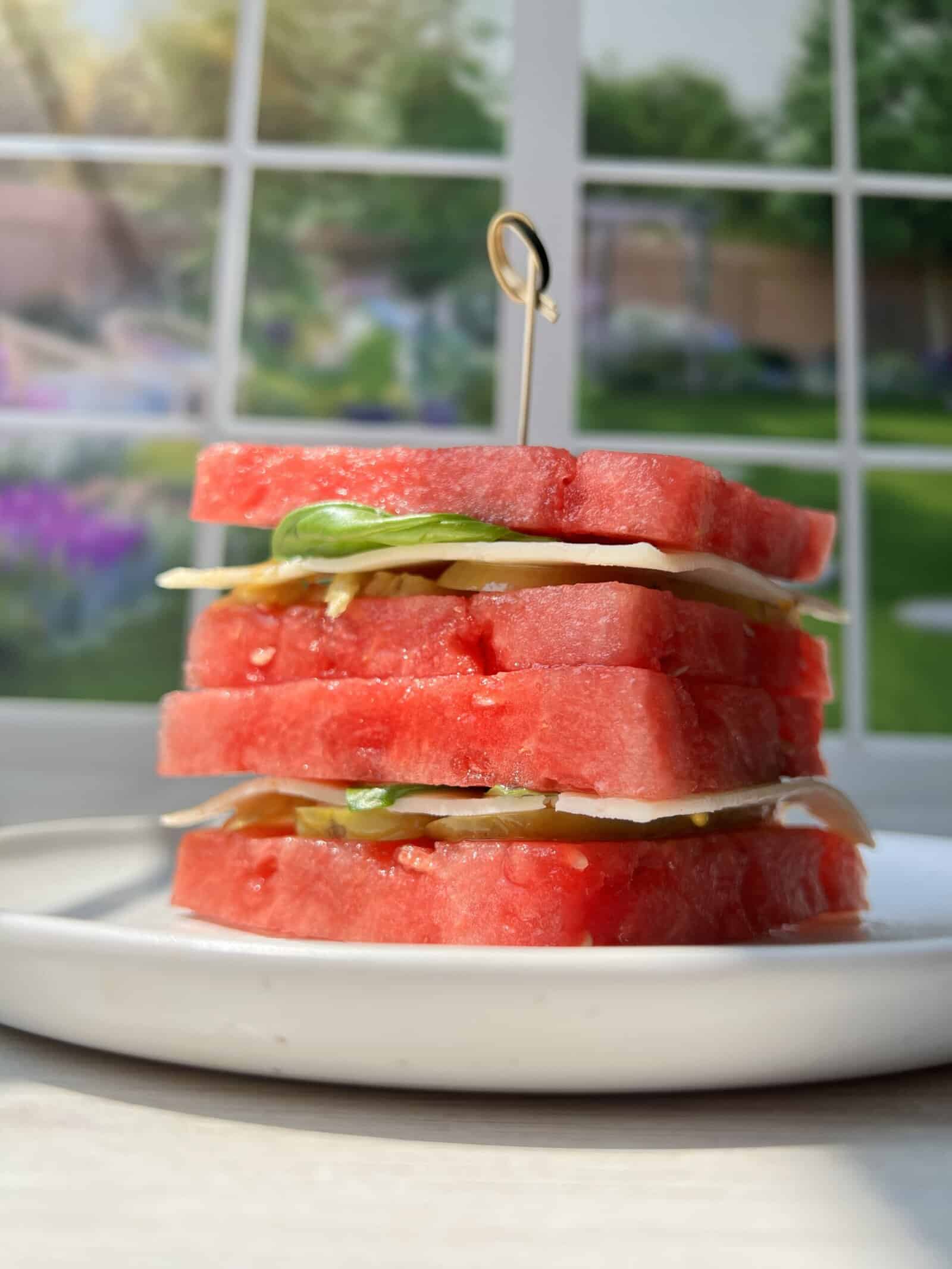 A stack of watermelon slices layered with mozzarella cheese and basil, secured with a wooden pick, sits on a white plate in front of a sunlit window with a garden view.