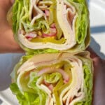 Two hands holding a lettuce wrap filled with slices of turkey, cheese, pickles, and shredded lettuce, shown in a cross-section view. The wrap is green and fresh, without bread or a tortilla.