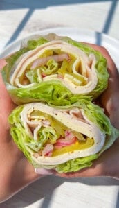 Two hands hold lettuce wraps filled with sliced turkey, cheese, pickles, onions, and bell peppers, shown in a close-up view with a white plate in the background.