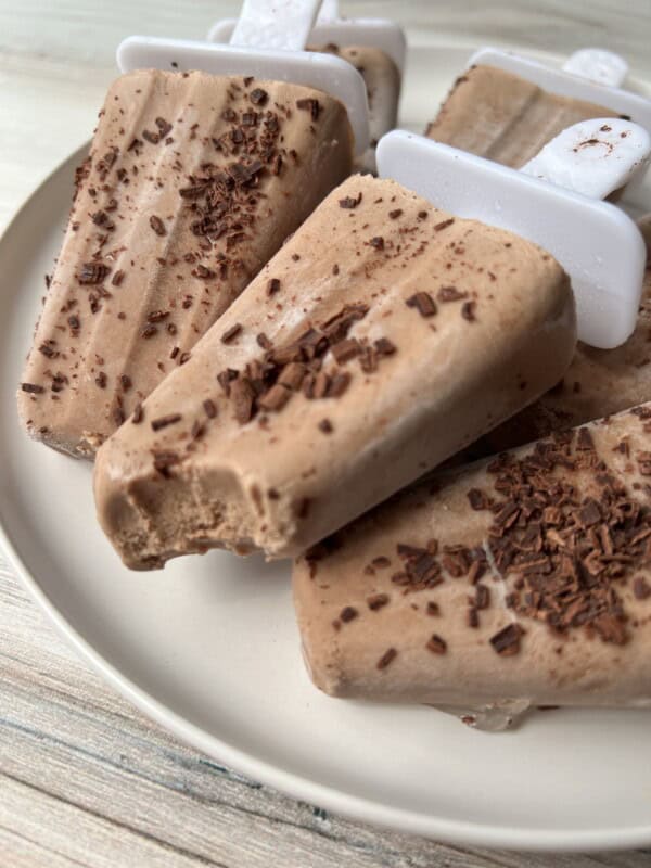A close-up of homemade chocolate popsicles on a plate, sprinkled with chocolate shavings. One popsicle has a visible bite taken out of it. Popsicle molds are still attached in the background.