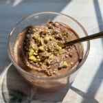 A clear glass filled with creamy chocolate pudding, topped with chopped nuts and seeds, sits on a sunlit wooden surface. A gold spoon is placed inside the glass. Shadows from window panes fall across the scene.