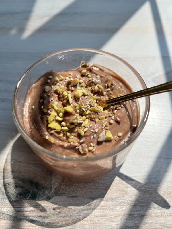 A clear glass filled with creamy chocolate pudding, topped with chopped nuts and seeds, sits on a sunlit wooden surface. A gold spoon is placed inside the glass. Shadows from window panes fall across the scene.