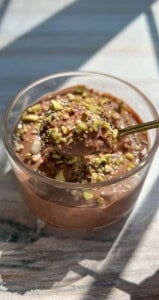 A glass cup filled with creamy chocolate mousse, topped with chopped pistachios. A spoon is lifting a portion of the mousse, showing its smooth texture. Sunlight illuminates the dessert on a wooden surface.