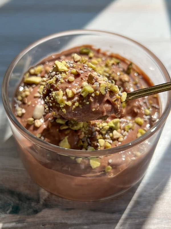 A glass cup filled with creamy chocolate mousse, topped with chopped pistachios. A spoon is lifting a portion of the mousse, showing its smooth texture. Sunlight illuminates the dessert on a wooden surface. - 6