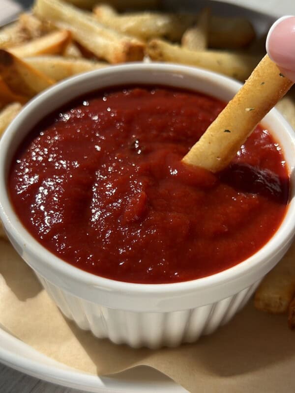 A French fry dipped into a small white ramekin filled with red ketchup, with more fries visible in the background on a plate.