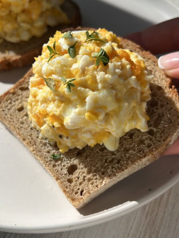 A close-up of a slice of brown bread topped with a scoop of creamy egg salad, garnished with fresh green herbs, held above a white plate in bright natural light.