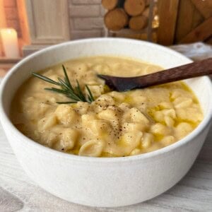 A bowl of creamy pasta with potatoes, garnished with black pepper and a sprig of rosemary, sits on a light wooden surface with a wooden spoon. The cozy background includes stacked firewood and a burning candle.