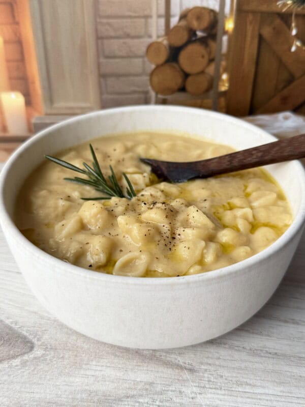 A bowl of creamy pasta with potatoes, garnished with black pepper and a sprig of rosemary, sits on a light wooden surface with a wooden spoon. The cozy background includes stacked firewood and a burning candle.