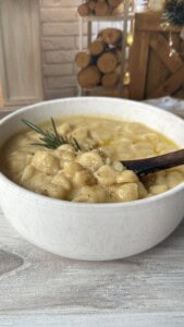 A creamy soup with gnocchi, garnished with a sprig of rosemary, served in a white bowl with a wooden spoon on a light wooden surface. Logs and rustic decor are blurred in the background.