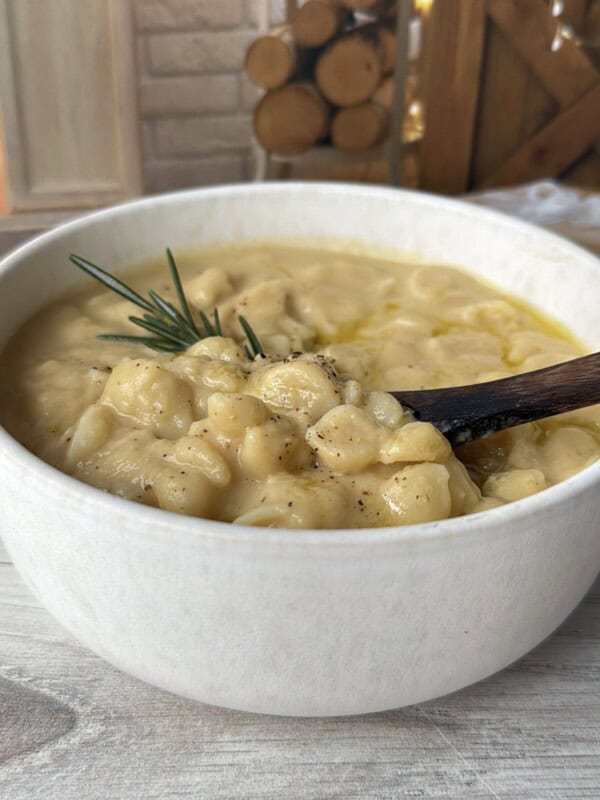 A creamy soup with gnocchi, garnished with a sprig of rosemary, served in a white bowl with a wooden spoon on a light wooden surface. Logs and rustic decor are blurred in the background. - 8