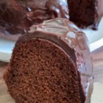 A close-up of a thick slice of chocolate Bundt cake with glossy chocolate glaze, placed on a wooden surface. The rest of the cake is visible on a white plate in the background.