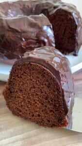 A close-up of a thick slice of chocolate Bundt cake with glossy chocolate glaze, placed on a wooden surface. The rest of the cake is visible on a white plate in the background.