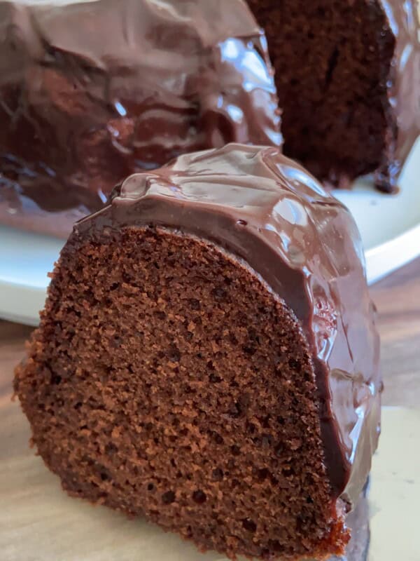 A close-up of a thick slice of chocolate Bundt cake with glossy chocolate glaze, placed on a wooden surface. The rest of the cake is visible on a white plate in the background.