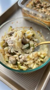 A close-up of a spoonful of creamy orzo pasta with chunks of chicken and zucchini, held above a glass bowl of the same dish. A baking dish with more of the casserole is in the background.