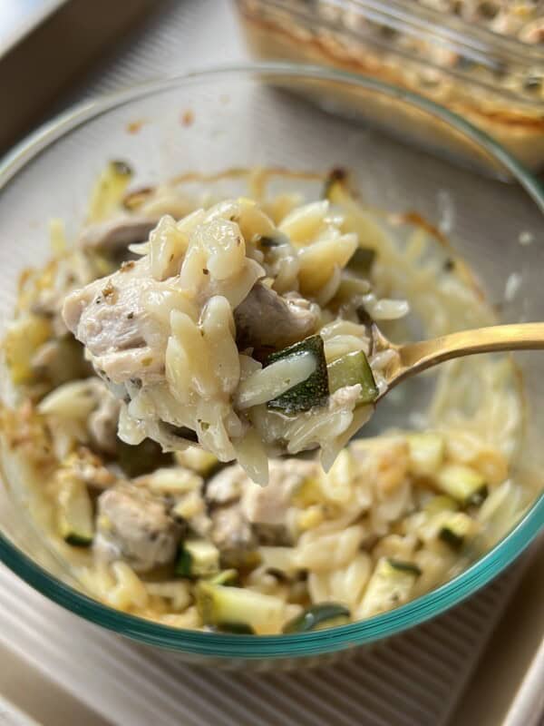 A close-up of a spoonful of creamy orzo pasta with chunks of chicken and zucchini, held above a glass bowl of the same dish. A baking dish with more of the casserole is in the background. - 11