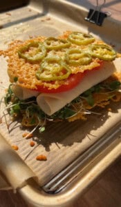 A sandwich with layers of microgreens, turkey slices, tomato, and crispy cheese topped with sliced jalapeños, served on a baking tray lined with parchment paper.