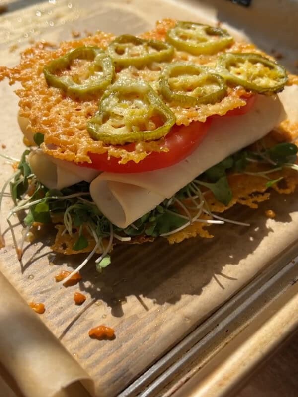 A sandwich with layers of microgreens, turkey slices, tomato, and crispy cheese topped with sliced jalapeños, served on a baking tray lined with parchment paper.