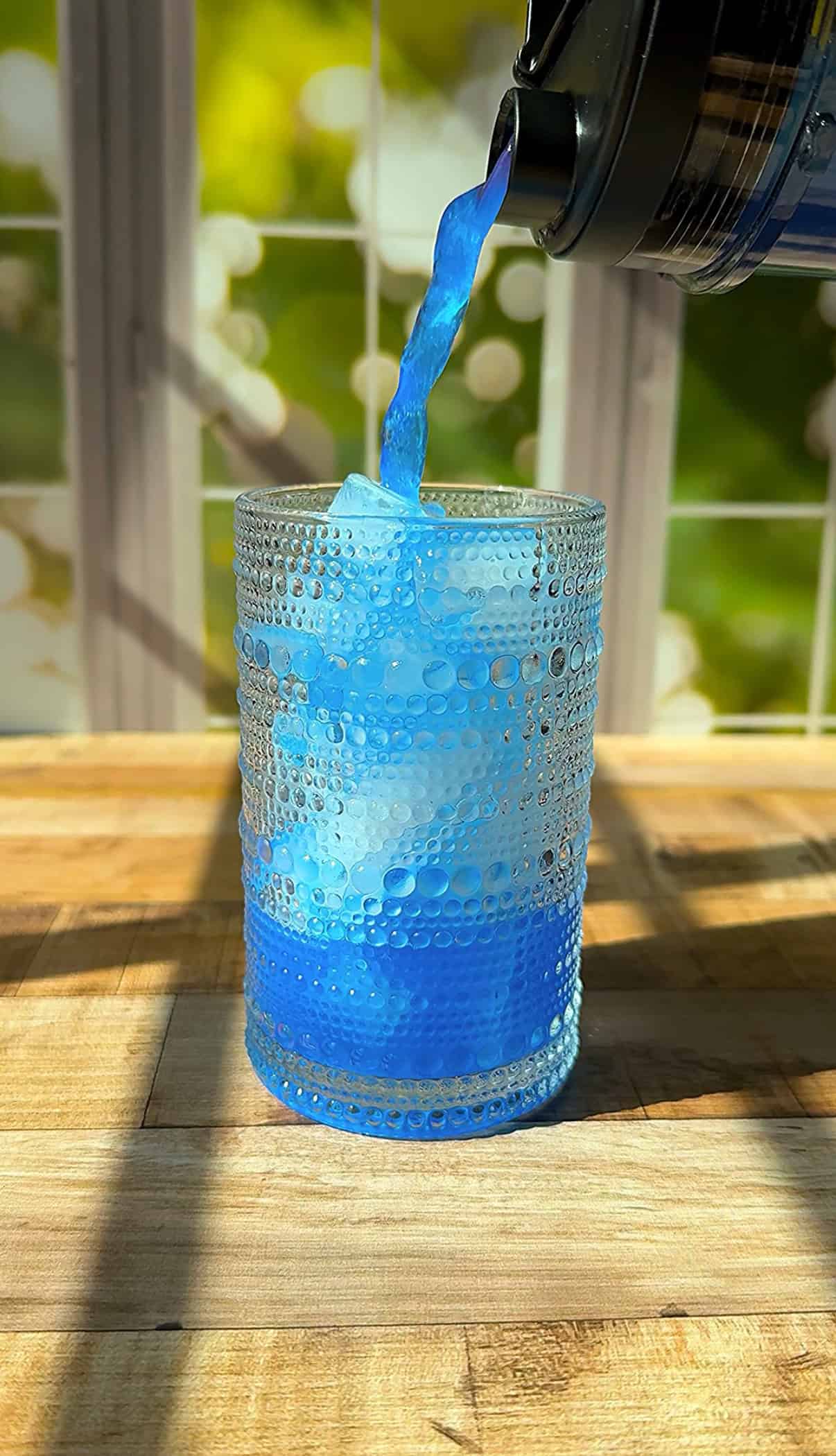 A bright blue liquid is being poured into a textured glass with ice cubes, set on a wooden surface near a sunlit window with a green, leafy background. - 3