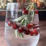 A clear glass of sparkling water with ice, garnished with fresh cranberries and rosemary sprigs, sits on a brown surface with a blurred festive background.