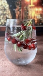 A clear glass of sparkling water with ice, garnished with fresh cranberries and rosemary sprigs, sits on a brown surface with a blurred festive background.