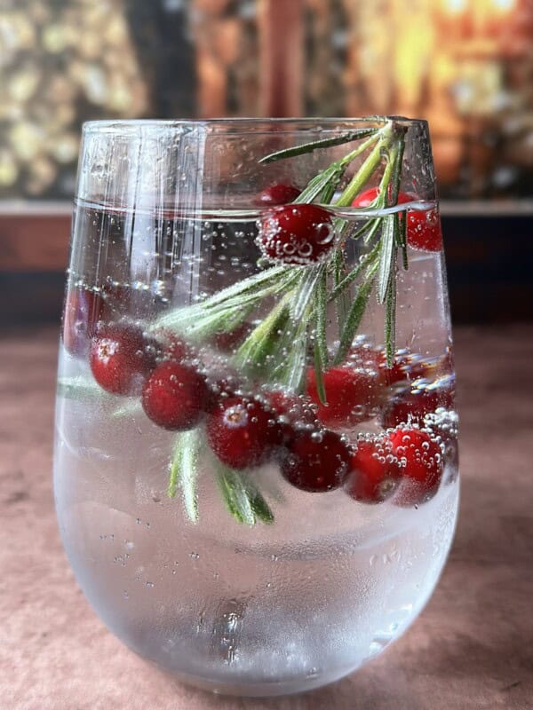 A clear glass of sparkling water with ice, garnished with fresh cranberries and rosemary sprigs, sits on a brown surface with a blurred festive background.