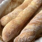 Three freshly baked baguettes with a golden, crispy crust rest on parchment paper in a white scalloped-edge baking dish.