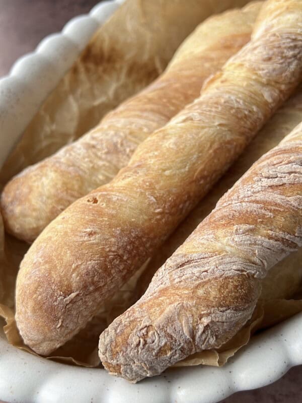 Three freshly baked baguettes with a golden, crispy crust rest on parchment paper in a white scalloped-edge baking dish.