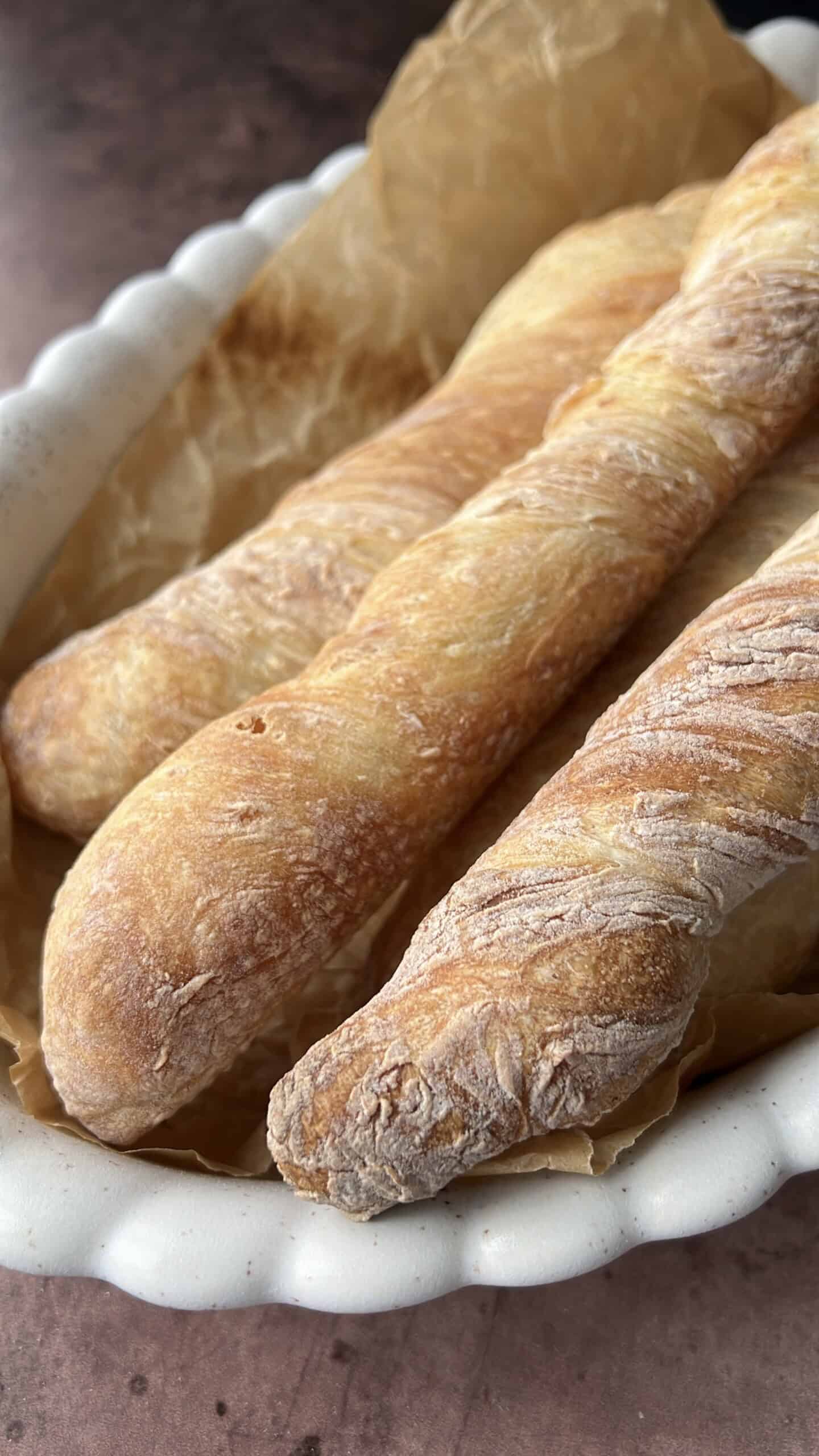 Three freshly baked baguettes with a golden, crispy crust rest on parchment paper in a white scalloped-edge baking dish. - 3