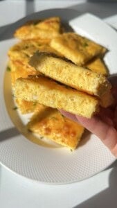 A hand holds two pieces of golden, fluffy baked egg squares with herbs, while more pieces are arranged on a textured white plate in bright sunlight.