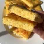 A hand holds two stacked, rectangular slices of fluffy, golden egg casserole with herbs, on a white plate with more pieces in the background, bathed in natural sunlight.