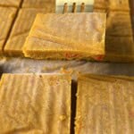 A metal fork lifts a square piece of golden-yellow fudge with red bits inside. More fudge squares are arranged on parchment paper in the background.