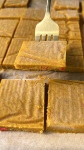 A metal fork lifts a square piece of golden-yellow fudge with red bits inside. More fudge squares are arranged on parchment paper in the background.