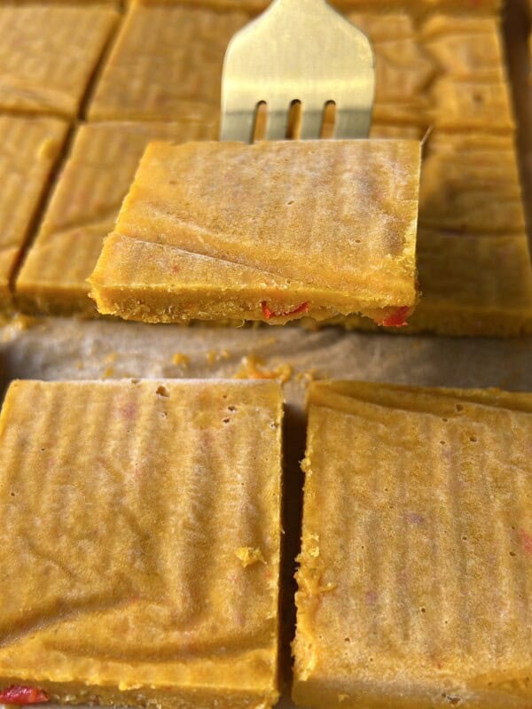 A metal fork lifts a square piece of golden-yellow fudge with red bits inside. More fudge squares are arranged on parchment paper in the background.