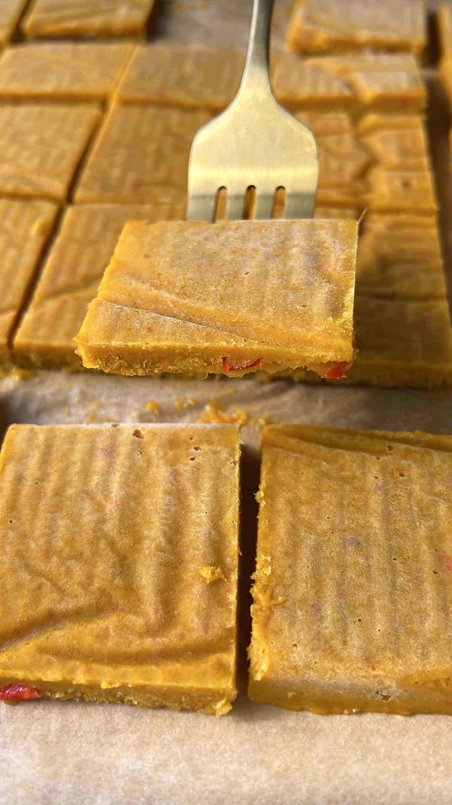 A metal fork lifts a square piece of golden-yellow fudge with red bits inside. More fudge squares are arranged on parchment paper in the background. - 3
