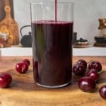 A glass is being filled with dark red cherry juice, placed on a wooden board with several fresh cherries around it. The background shows kitchen items on a counter.