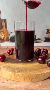 A glass is being filled with dark red cherry juice, placed on a wooden board with several fresh cherries around it. The background shows kitchen items on a counter.