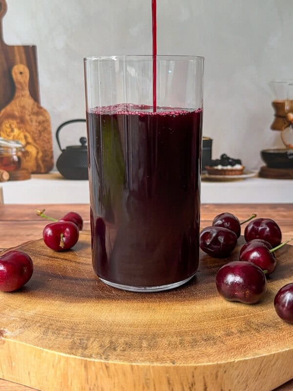 A glass is being filled with dark red cherry juice, placed on a wooden board with several fresh cherries around it. The background shows kitchen items on a counter.