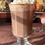 A glass mug of hot chocolate topped with whipped cream and gold sprinkles sits on a sunlit surface, with a blurred festive background.