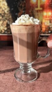 A glass mug of hot chocolate topped with whipped cream and gold sprinkles sits on a sunlit surface, with a blurred festive background.