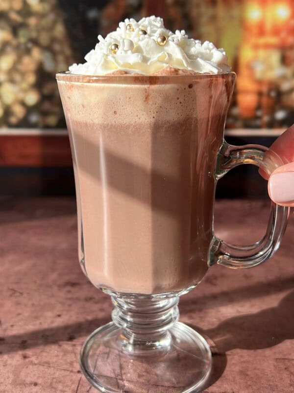 A glass mug of hot chocolate topped with whipped cream and gold sprinkles sits on a sunlit surface, with a blurred festive background.