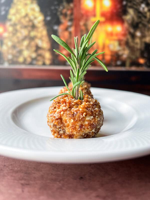 A close-up of a round, breaded appetizer on a white plate, garnished with a sprig of fresh rosemary. The background is softly blurred with warm, festive lights.