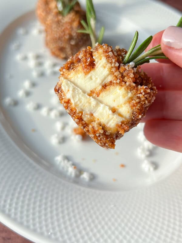A hand holds a piece of golden-brown, breaded cheese on a sprig of rosemary, showing its soft inside. Two more pieces are on a white plate with scattered cheese crumbles.