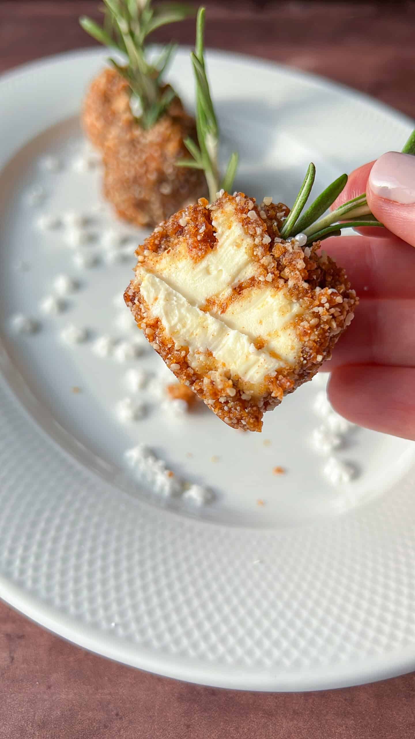 A hand holds a piece of golden-brown, breaded cheese on a sprig of rosemary, showing its soft inside. Two more pieces are on a white plate with scattered cheese crumbles.
