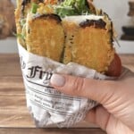 A hand holds a sandwich wrapped in newspaper, filled with golden fried zucchini slices, fresh greens, and cheese, on a wooden table.
