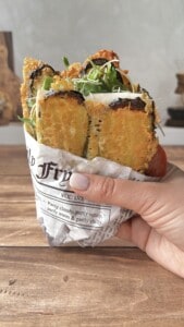 A hand holds a sandwich wrapped in newspaper, filled with golden fried zucchini slices, fresh greens, and cheese, on a wooden table.