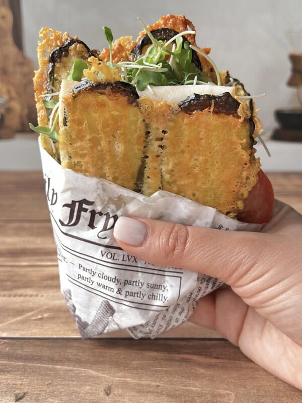 A hand holds a sandwich wrapped in newspaper, filled with golden fried zucchini slices, fresh greens, and cheese, on a wooden table.